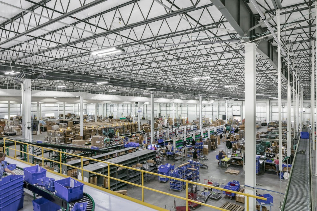 Wide interior view of a large industrial warehouse with high steel truss ceilings, assembly lines, stacked boxes, blue storage bins, and workers operating at packing and distribution stations.